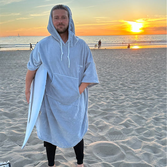Man wearing the Venice Surf Club hooded surf poncho in Shark Gray, holding a surfboard on the beach at sunset